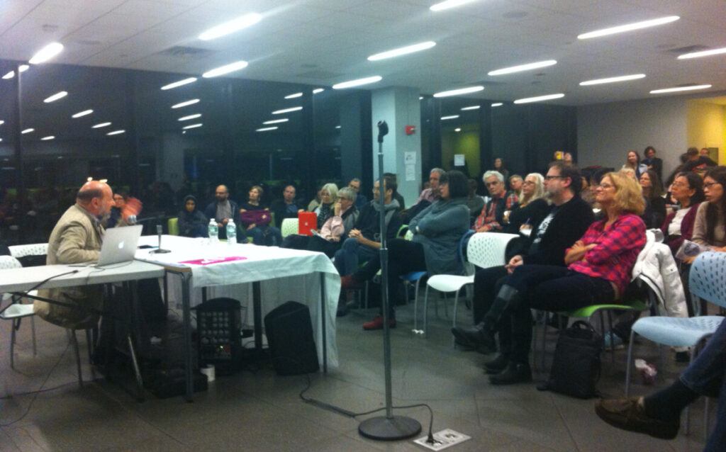 A speaker addresses an audience seated in a bright room with large windows. A table with a laptop is in front of the speaker. Attendees appear engaged, sitting on white chairs. 